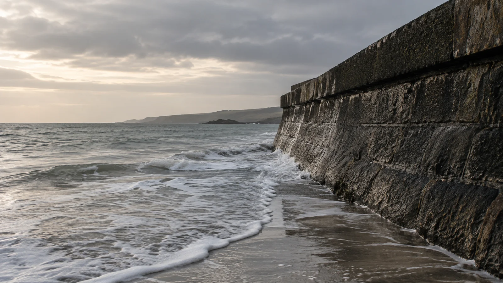 Conceptual photograph: A receding wave against a sturdy dark stone seawall, quiet overcast coast, not a storm