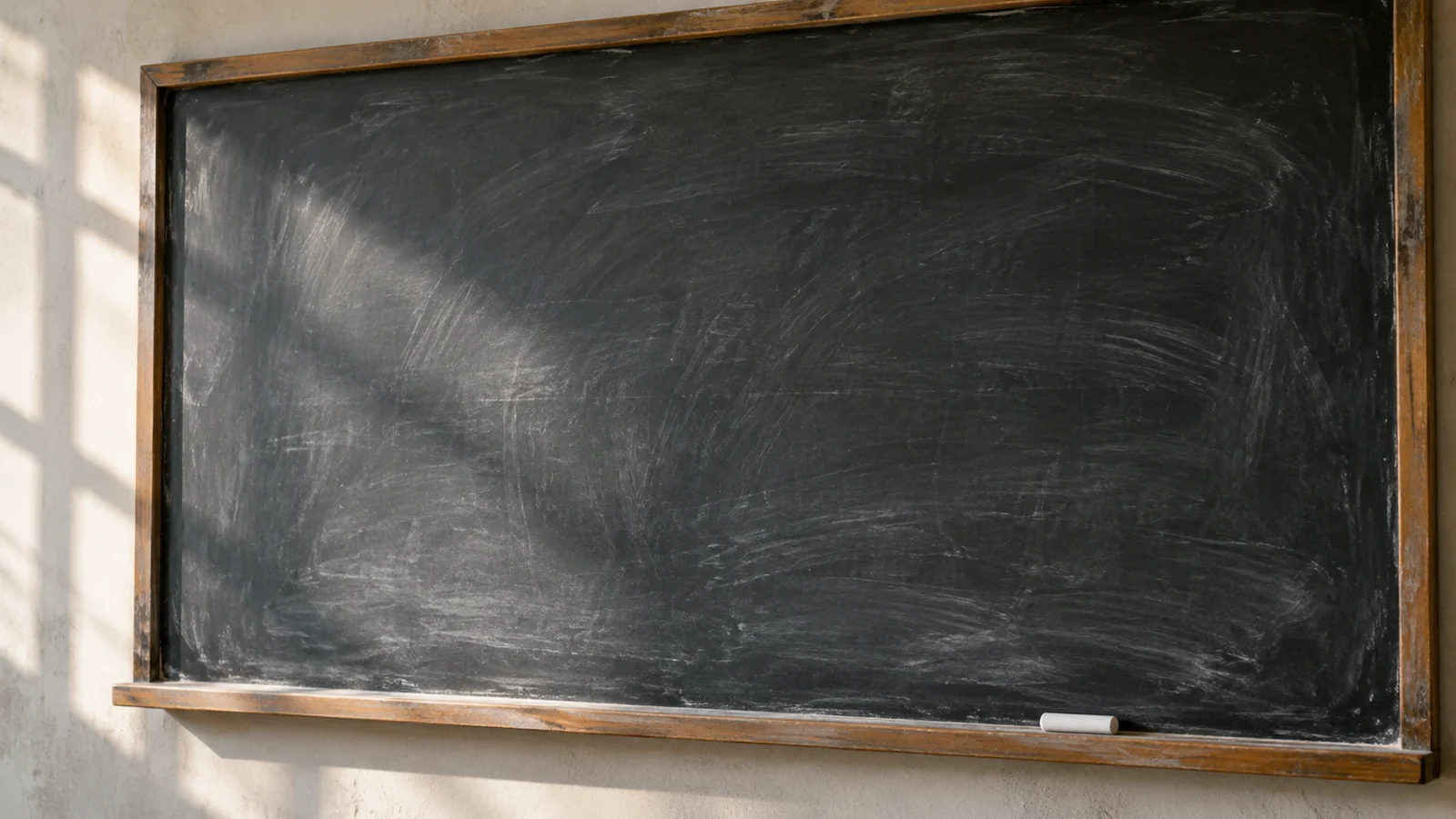 Conceptual photograph: A clean slate blackboard with a few erased chalk traces and one piece of chalk on the ledge, no writing